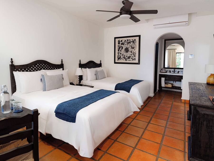 Habitación de hotel con dos camas, un ventilador de techo, una obra de arte enmarcada, un baño visible a través de un arco y un televisor sobre un pedestal.