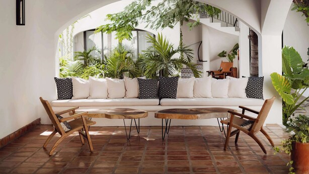 A modern living room with a white sofa, two wooden chairs, and a coffee table. There is a large window with plants and a view of a staircase. - Tropicana Los Cabos, Tapestry Collection by Hilton (San José del Cabo)
