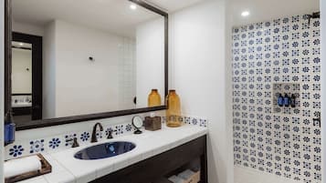 Un baño con un lavabo blanco, tocador de madera oscura y un espejo. Hay dos botellas en la encimera y un perchero para toallas.