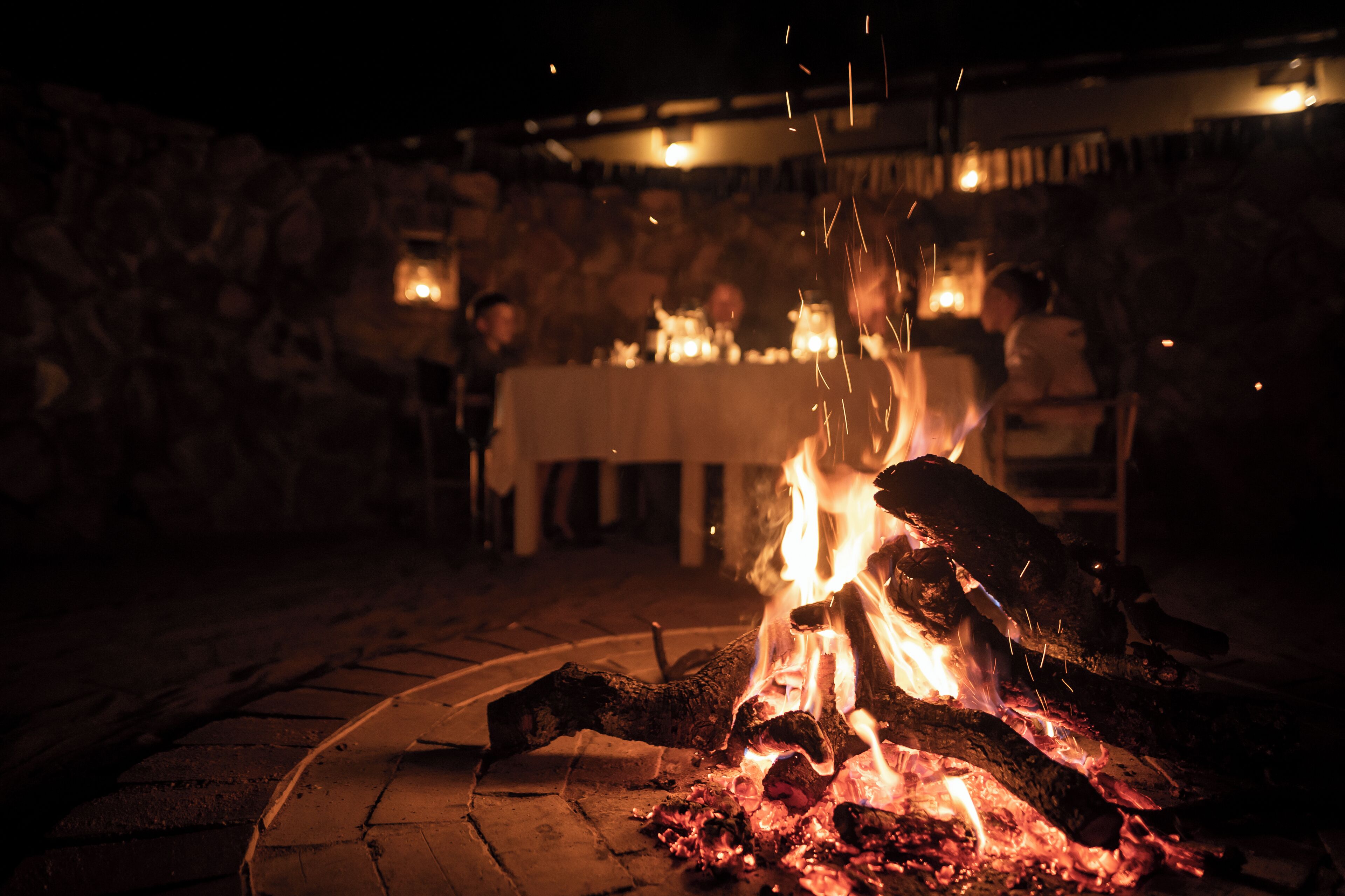 A fire pit with burning wood, surrounded by stone walls and tables with candles.