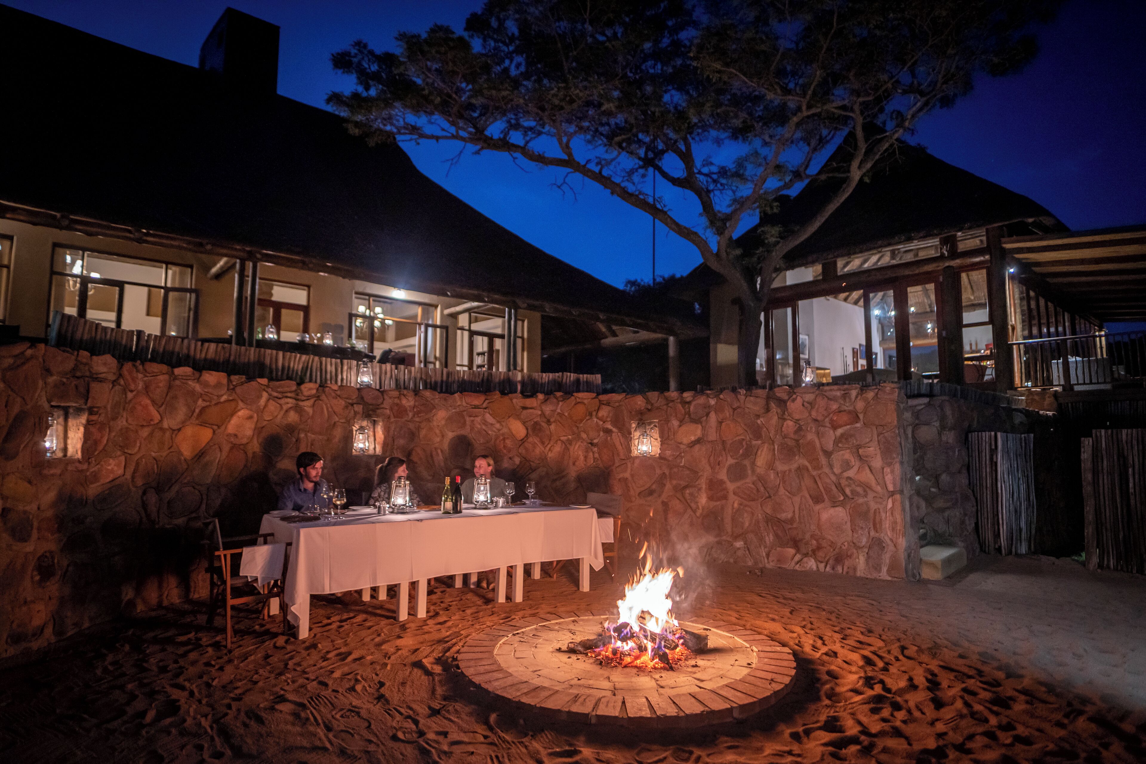 A stone-walled outdoor dining area with a fire pit, tables set for dining, and a view of the building's interior.
