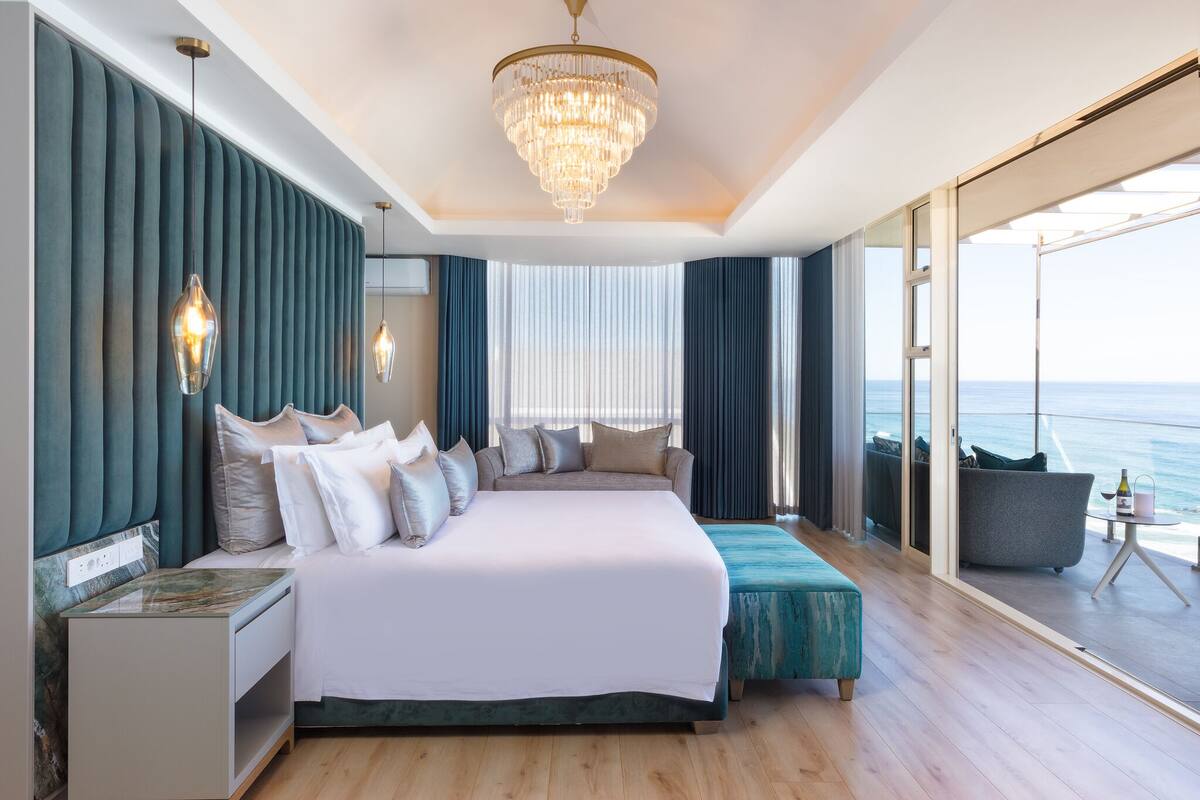 A spacious bedroom with a large bed, a chandelier, a view of the ocean, and a balcony with a table and chairs.