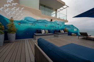 Una terraza con zonas de estar, un mural de aves y olas, y vista al mar.