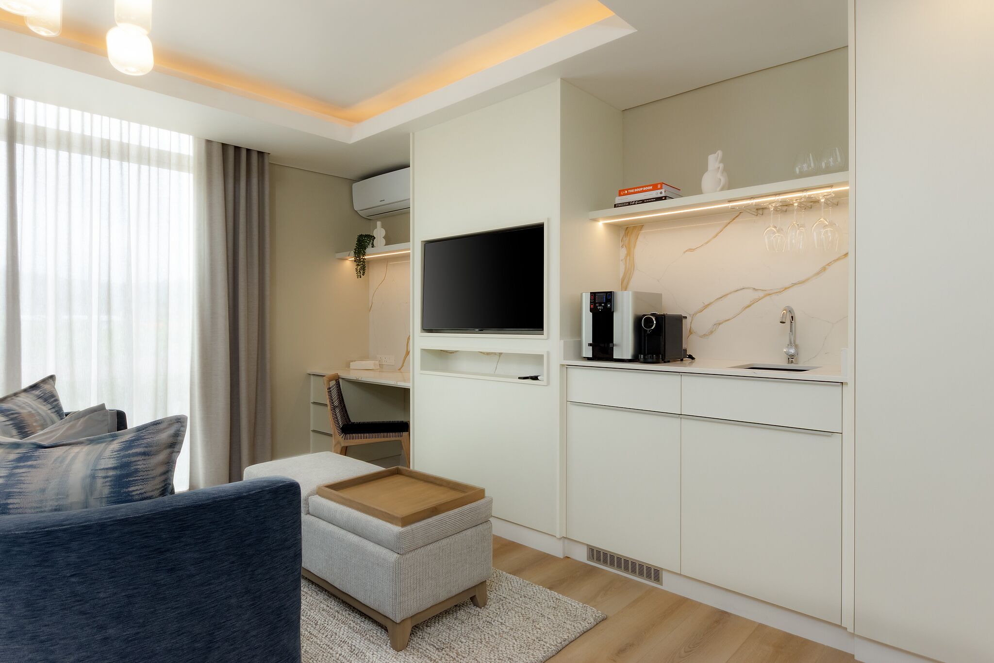 A modern living room with a sofa, ottoman, TV, and kitchen area with cabinets and a coffee machine.