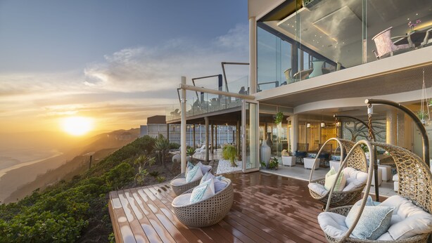 Een modern terras buiten met rieten stoelen en een glazen balustrade met uitzicht op een schilderachtig landschap.