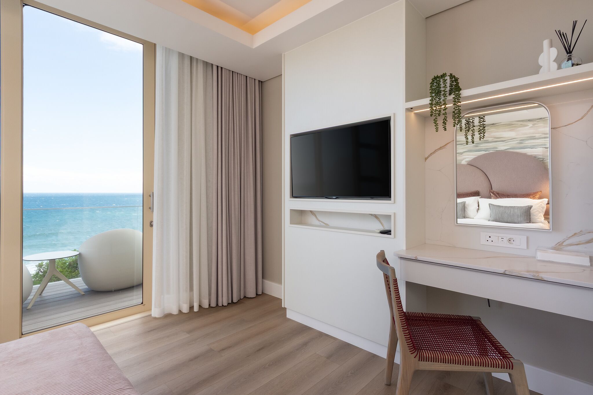 A modern hotel room with a large window, a flat-screen TV, a desk, and a chair.