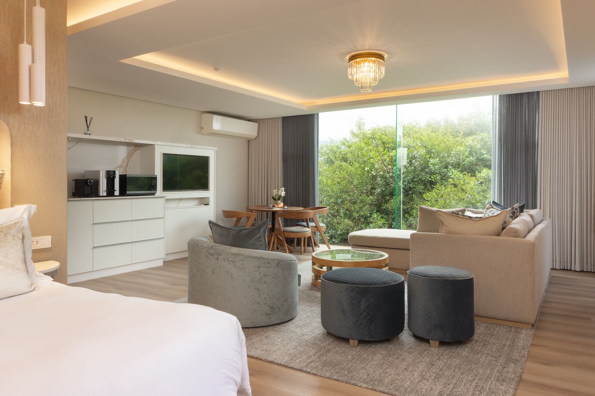 A modern hotel room with a bed, a sofa, ottomans, a dining table, and a TV.