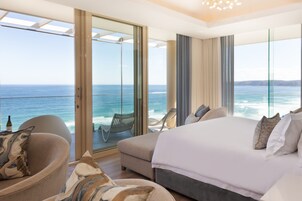 Quarto de hotel moderno com cama grande, área de estar e varanda com vista para o oceano.