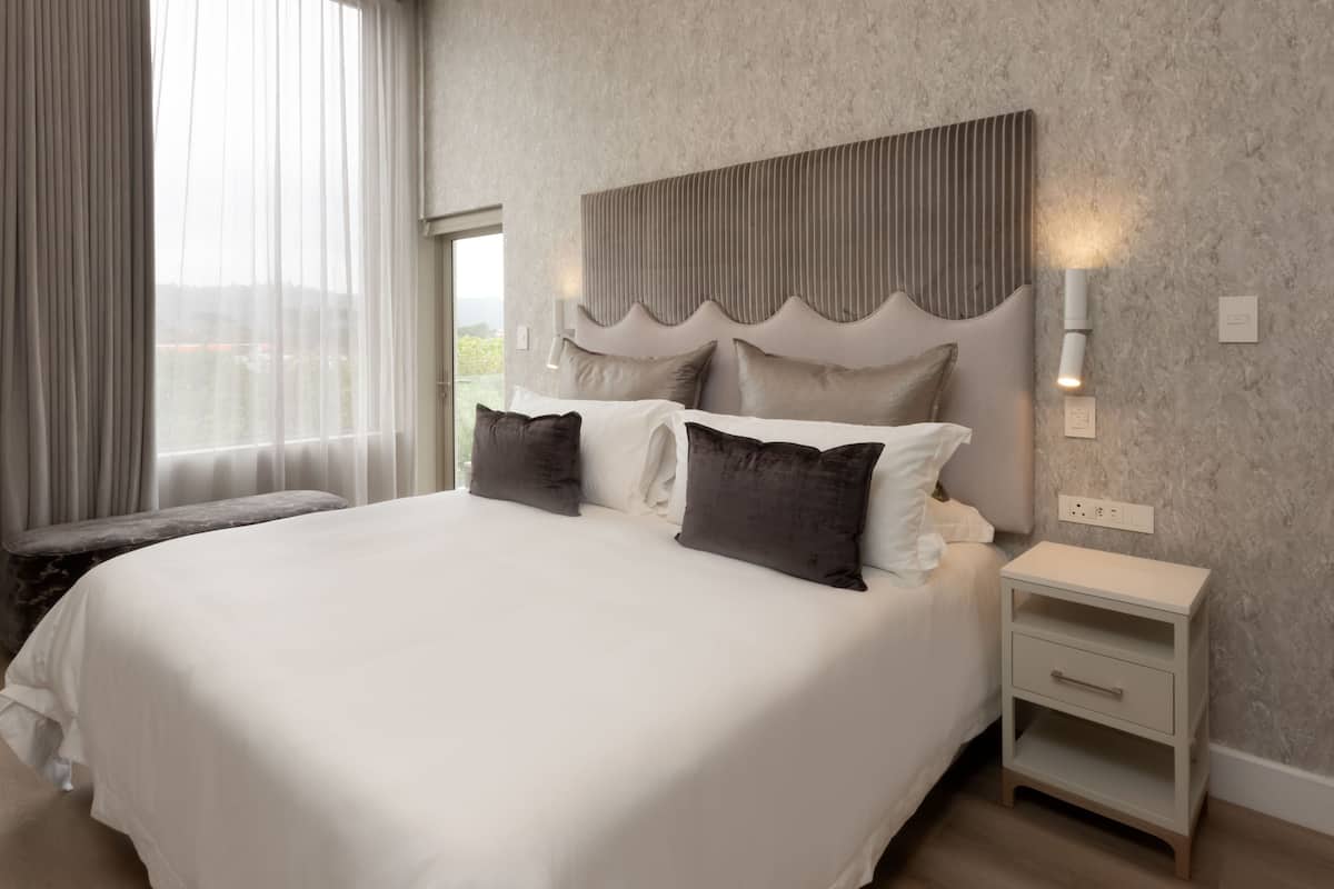 A modern hotel room with a large bed, a bedside table, and a view of the outdoors.