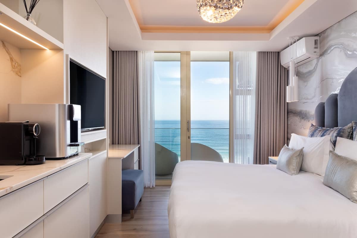 A modern hotel room with a large bed, a desk, a TV, and a view of the sea.