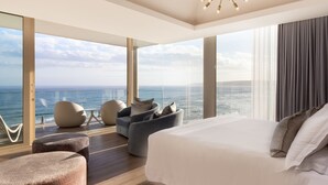 Een moderne slaapkamer met een groot bed, twee fauteuils, een klein voetenbankje en uitzicht op zee.