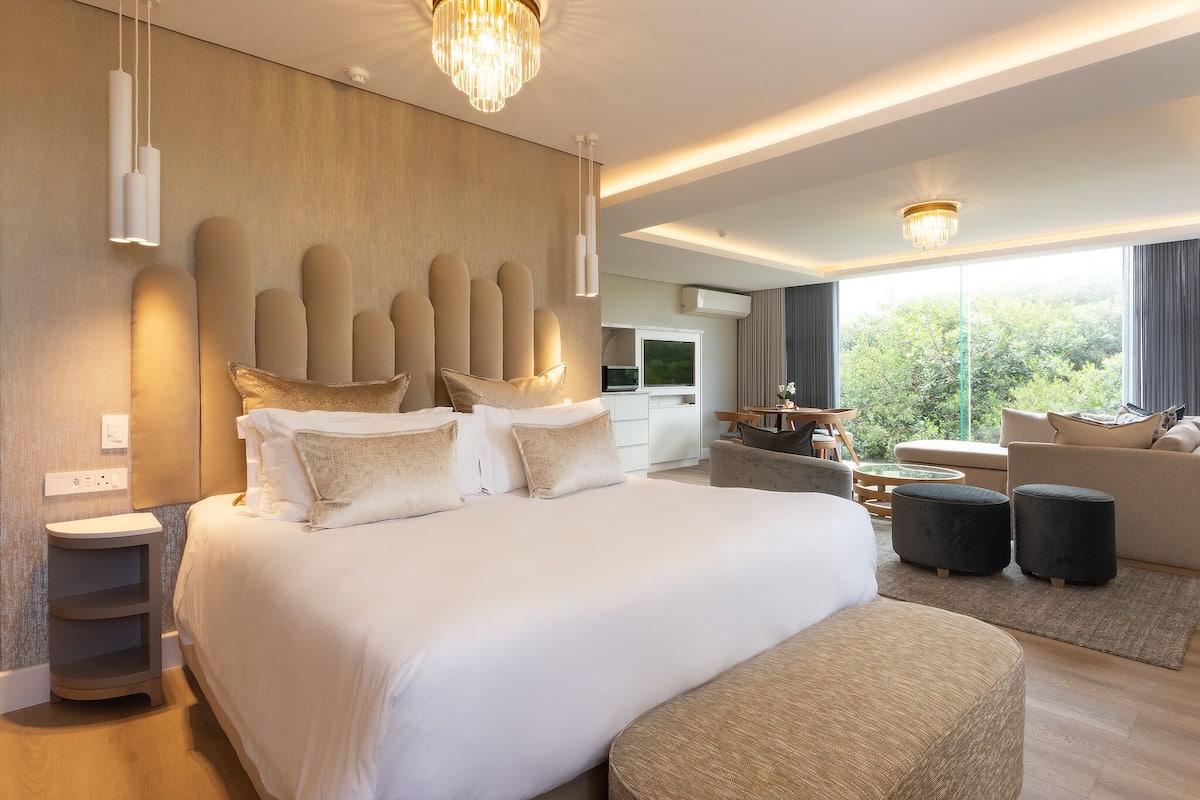 A modern hotel room with a large bed, bedside tables, a sofa, ottomans, a chandelier, and a view of greenery outside.