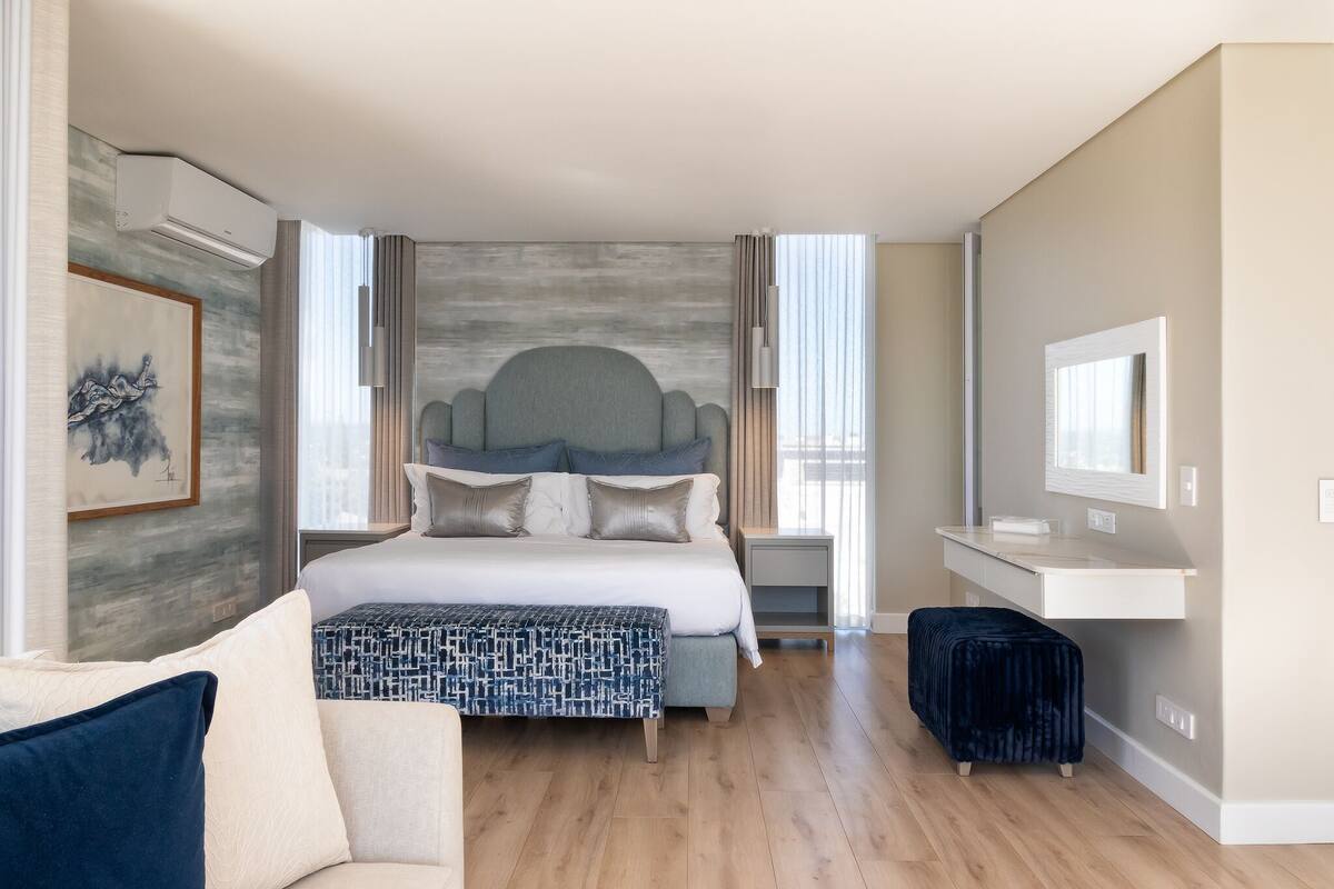 A modern hotel room with a large bed, a bedside table, a TV, and a seating area with a sofa and ottoman.