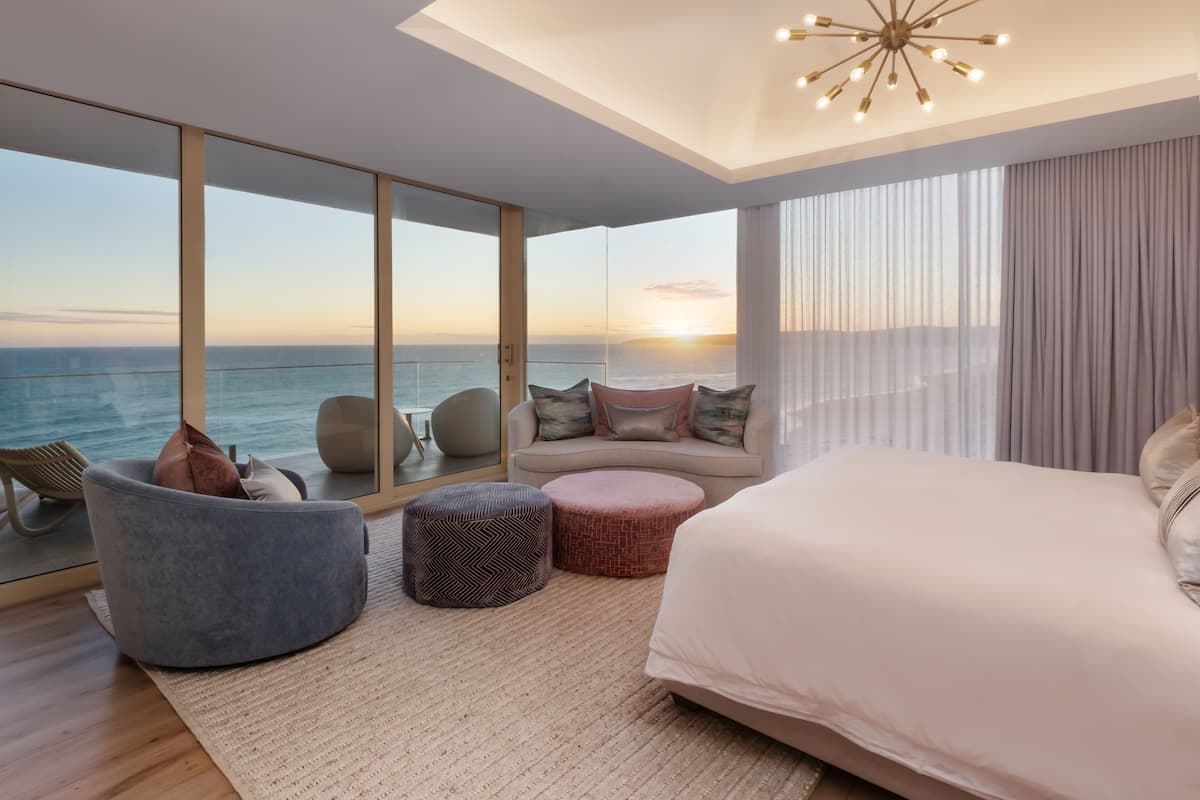 A modern bedroom with a large bed, a sofa, and a chair, featuring a view of the ocean and a sunset.