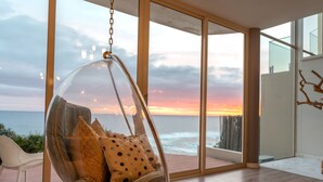 Een hangstoel met kussens in een kamer met grote ramen met uitzicht op de oceaan bij zonsondergang.