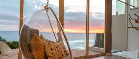 A hanging chair with cushions in a room with large windows overlooking the ocean at sunset.