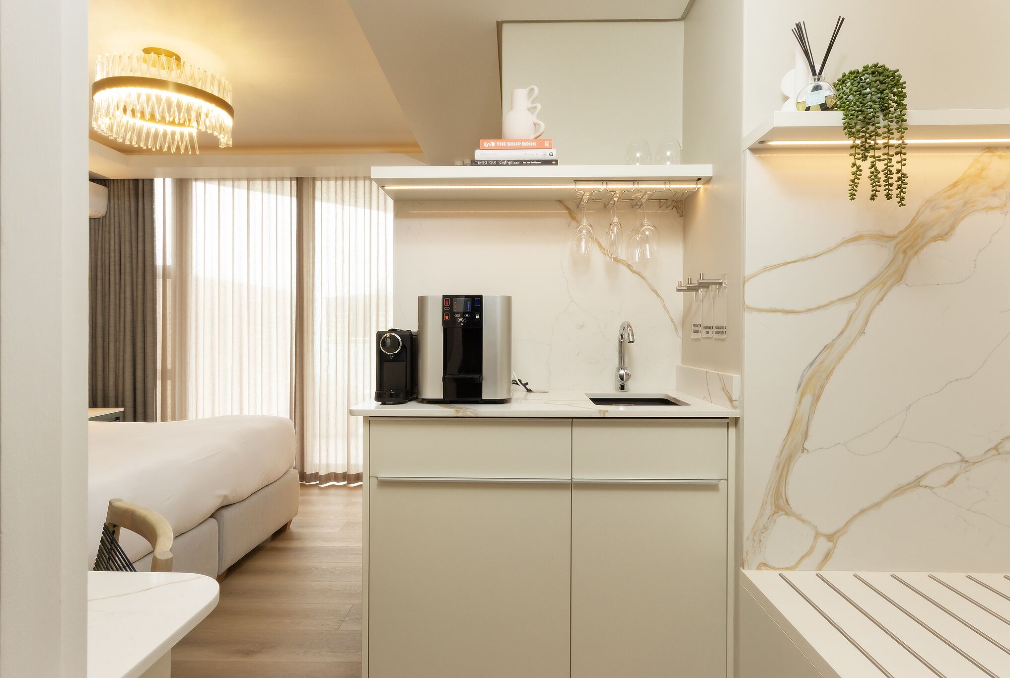A compact hotel room with a bed, a small kitchenette with a coffee maker, a mirror, and a marble wall.