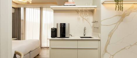 A compact hotel room with a bed, a small kitchenette with a coffee maker, a mirror, and a marble wall.