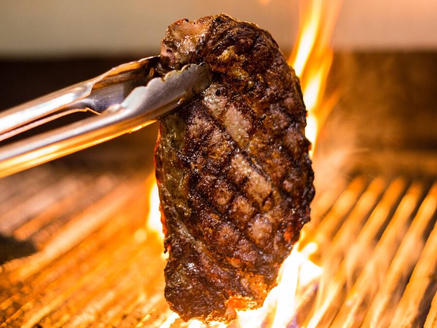 Gros plan d’un steak grillé, maintenu à l’aide de pinces au-dessus d’un gril.