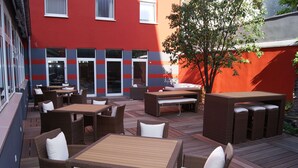 An outdoor patio with wicker furniture, a wooden table, and a red building in the background. - Cityhotel Thüringer Hof new CLASSIC (Hannover)