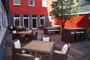 Un patio al aire libre con mobiliario de mimbre, una mesa de madera y un edificio rojo al fondo.