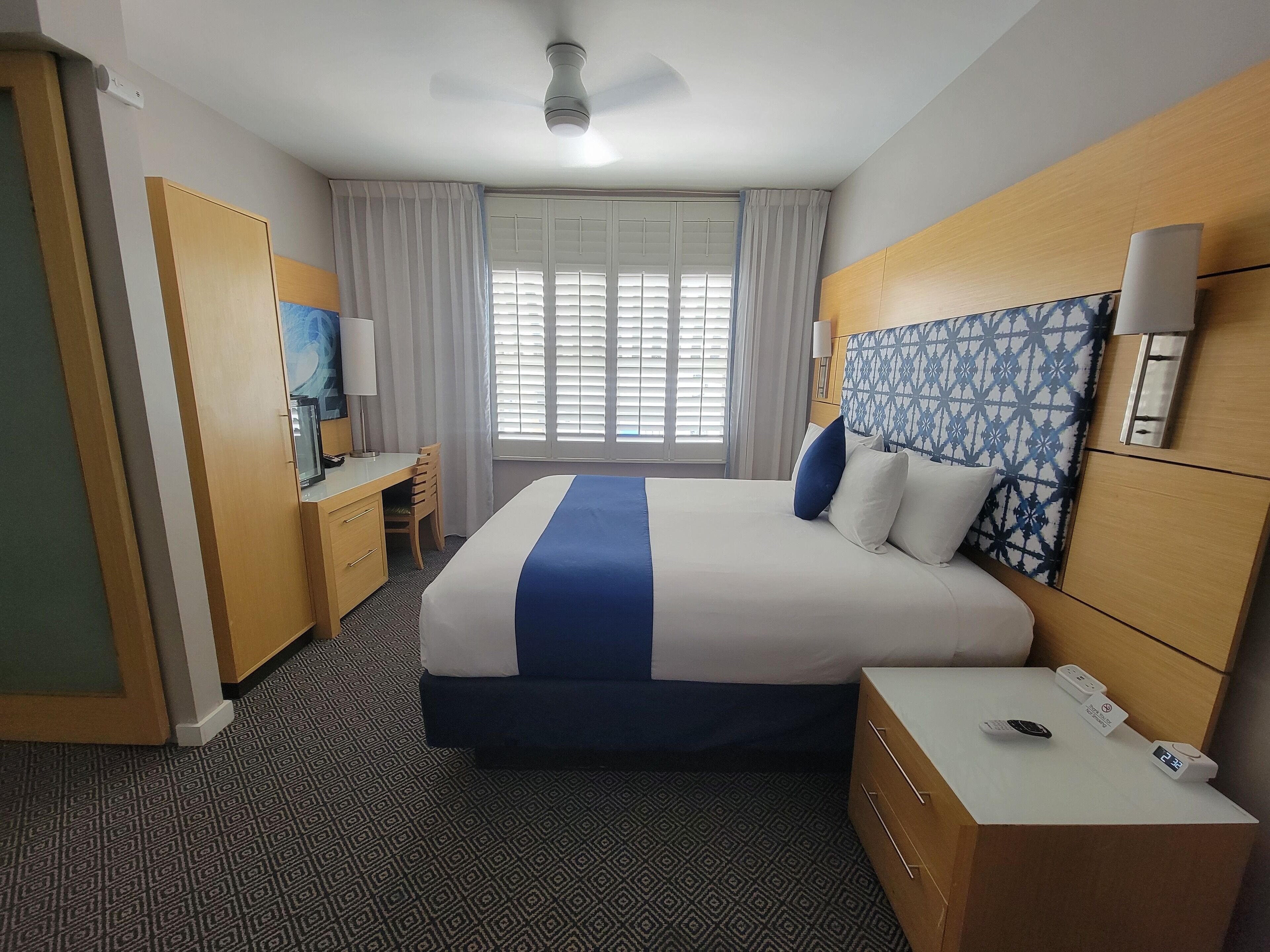 A hotel room with a bed, a desk with a chair, a nightstand with a phone, a wall-mounted lamp, and a window with blinds.