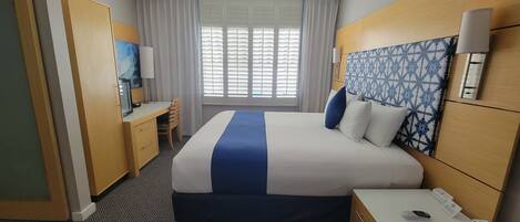 A hotel room with a bed, a desk with a chair, a nightstand with a phone, a wall-mounted lamp, and a window with blinds.