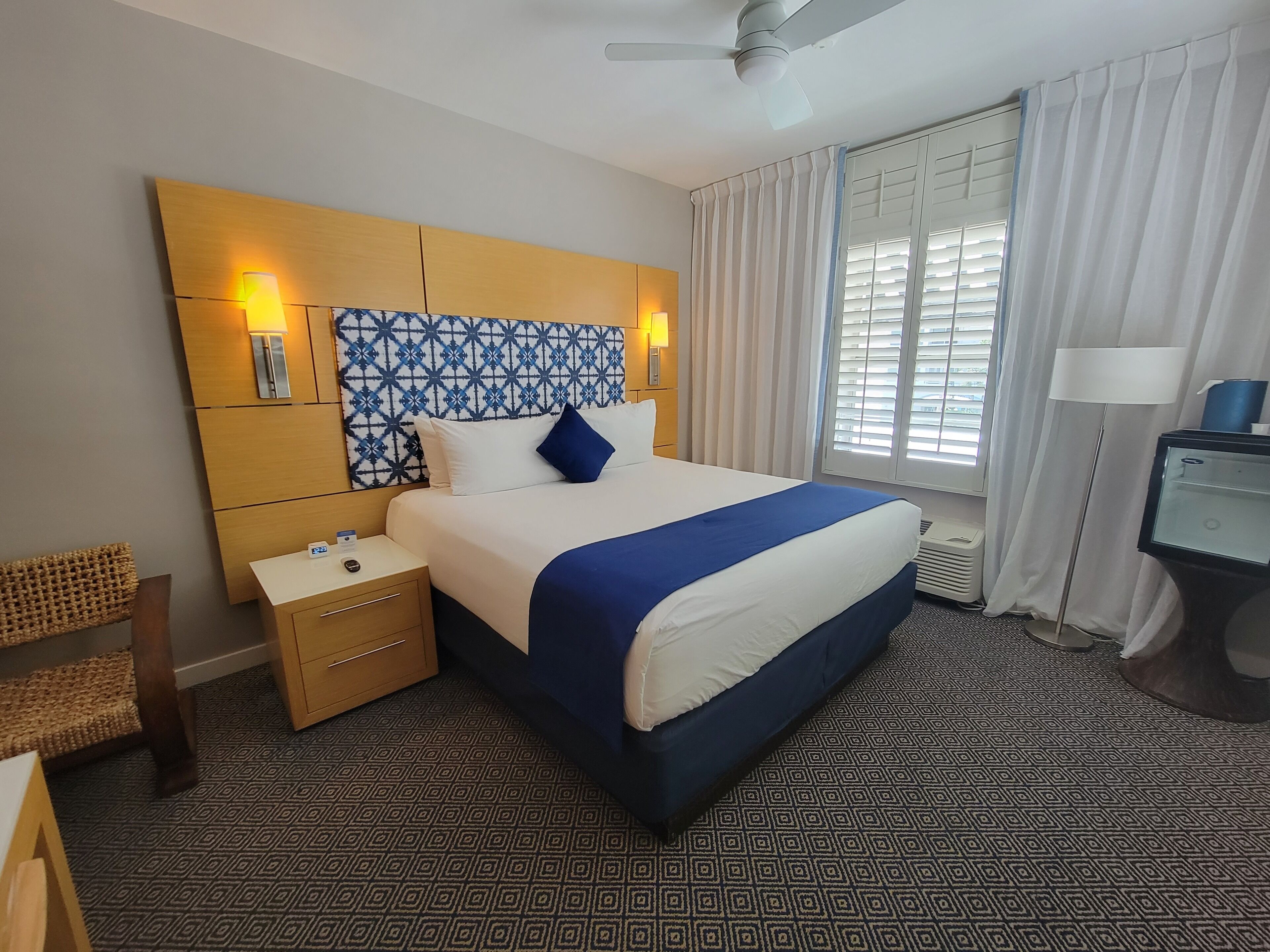 A hotel room with a bed, bedside tables, a desk, a chair, a ceiling fan, and a window with curtains.