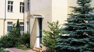 Ein Wohngebäude mit einem schmalen Weg zum Eingang, umgeben von Grünflächen und einem hohen Nadelbaum.