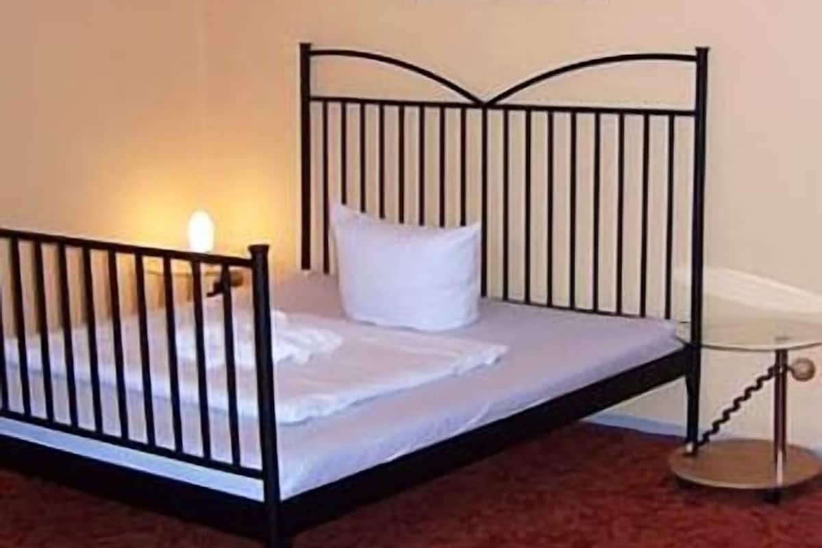A single bed with a black metal frame, white bedding, and a small bedside table with a lamp.
