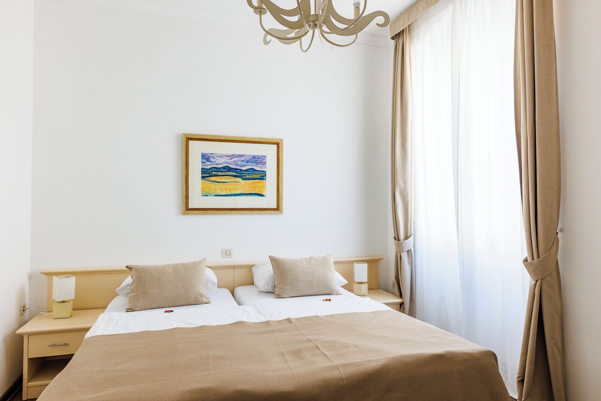 Habitación de hotel con cama, dos mesitas de noche, una lámpara, un cuadro enmarco y una ventana con cortinas.