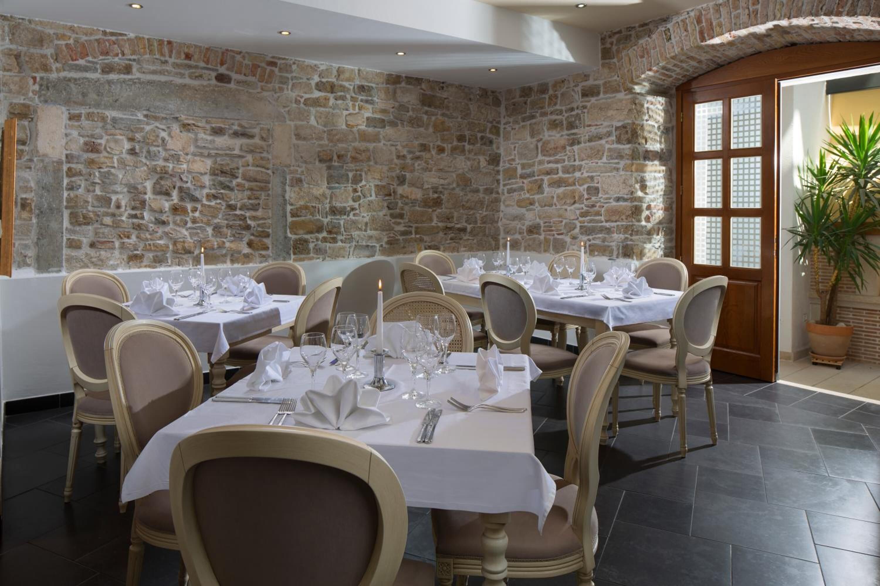 Un restaurant aux murs de pierre et à la porte en bois, avec des tables recouvertes de nappes blanches et décorées de bougies.