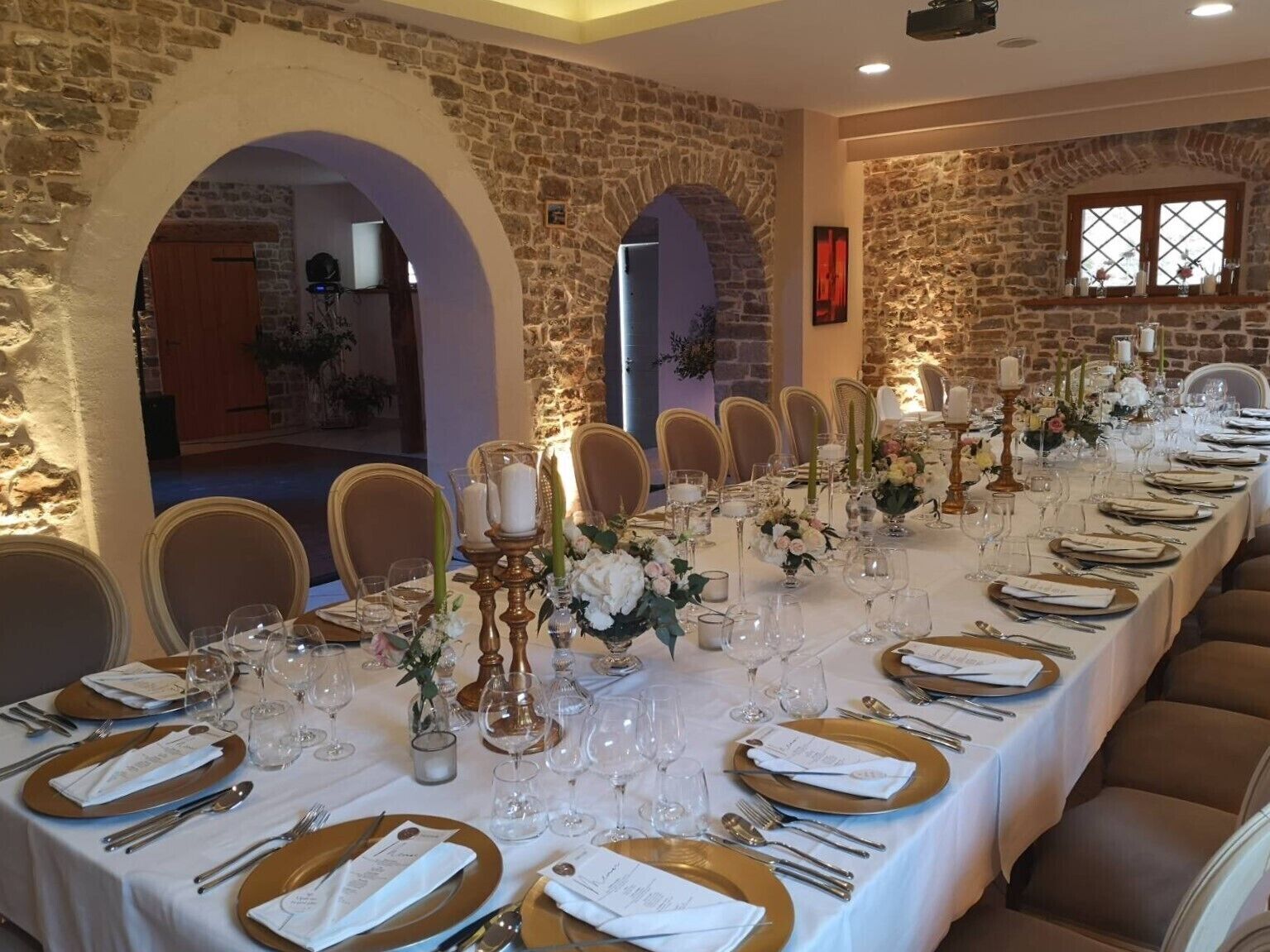 Una larga mesa de comedor dispuesta para un evento formal, con manteles blancos, platos base dorados y centros de mesa florales.