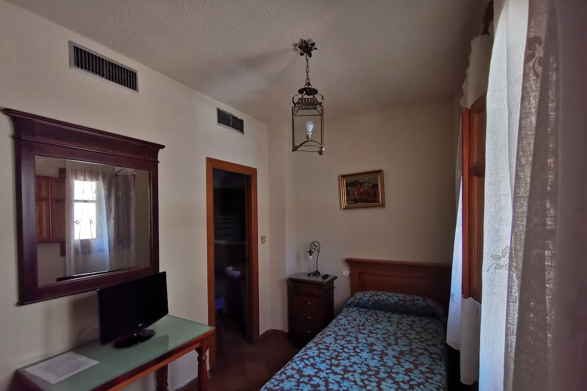 A bedroom with a bed, a desk with a computer, a mirror, a hanging light fixture, and a door leading to another room.