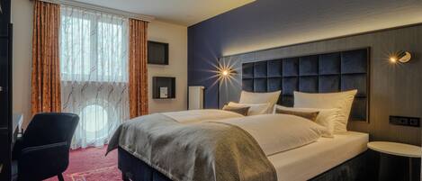 Habitación de hotel con una cama grande, un escritorio, una silla, una mesita y una ventana con cortinas.