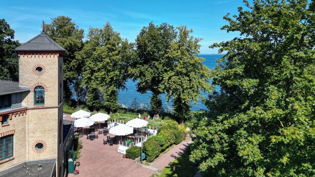 A building with a tower, outdoor seating area with umbrellas, and a view of the sea. - Hotel Seeblick Wismar (Wismar)