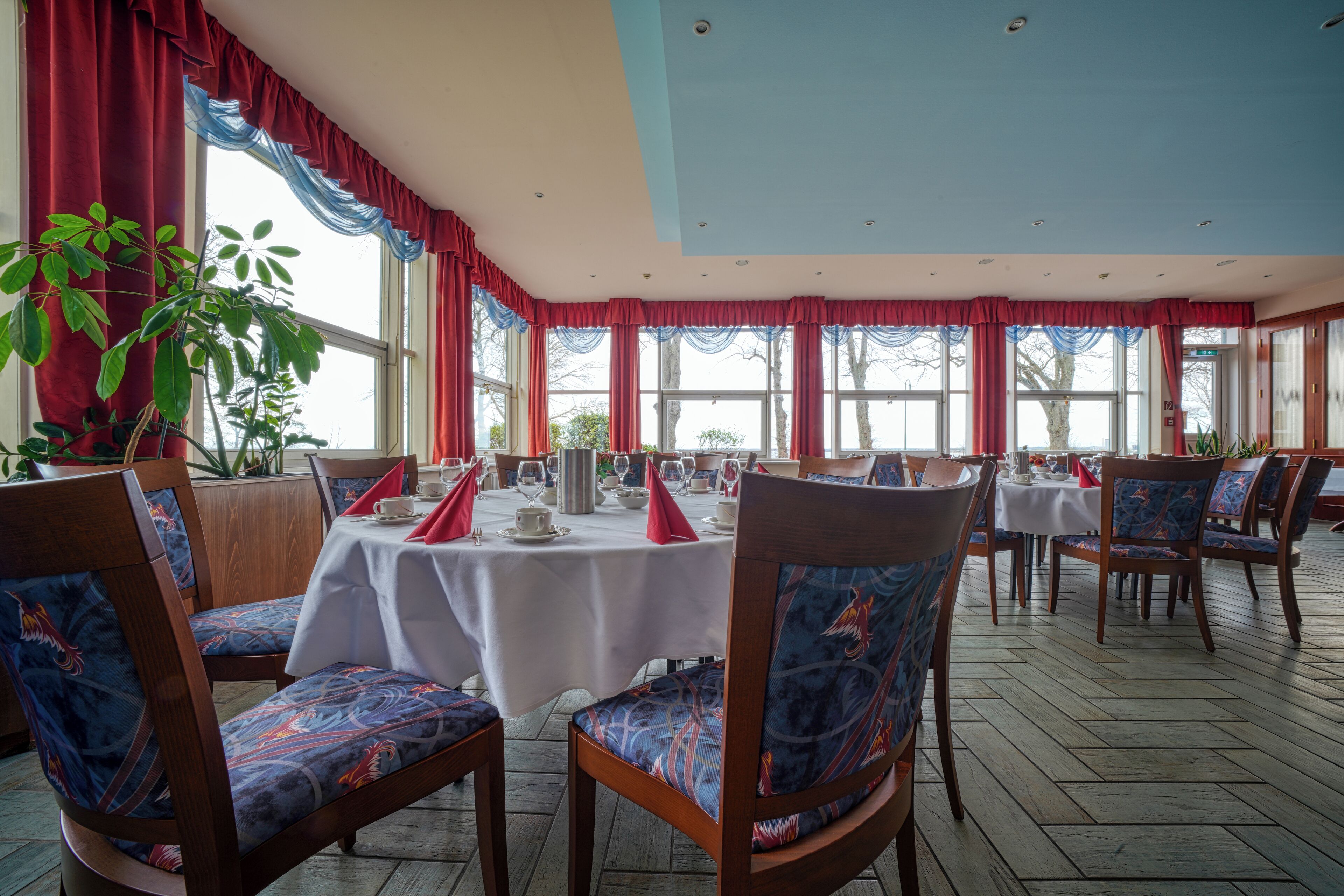 Un comedor con sillas y mesas de madera, ventanas amplias con cortinas rojas y plantas en macetas.