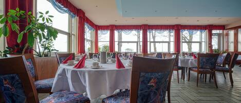 A dining area with wooden chairs and tables, large windows with red curtains, and potted plants.