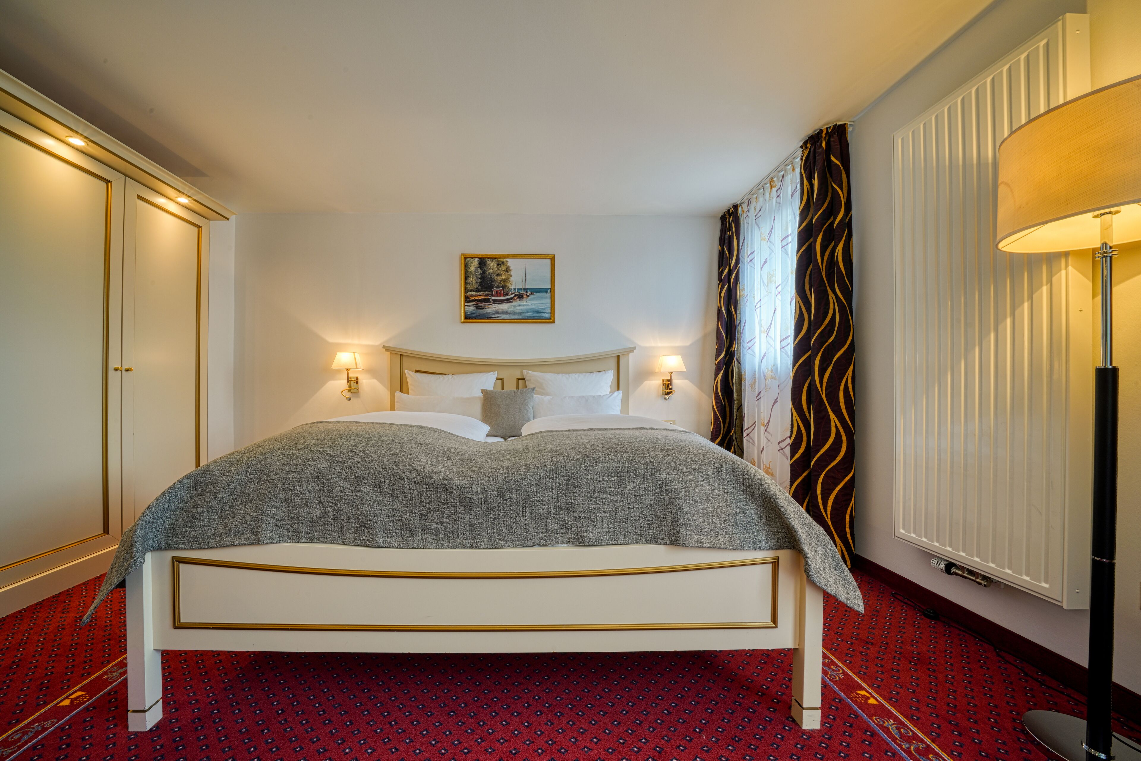 A neatly made bed with a white headboard and a grey blanket, a painting on the wall, and a red patterned carpet.