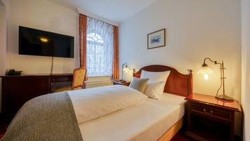Ein Hotelzimmer mit einem großen Bett, einem Schreibtisch mit Flachbildfernseher, einem Sessel, einem Fenster mit Vorhängen, einem Nachttisch mit Lampe und einem gerahmten Bild an der Wand.