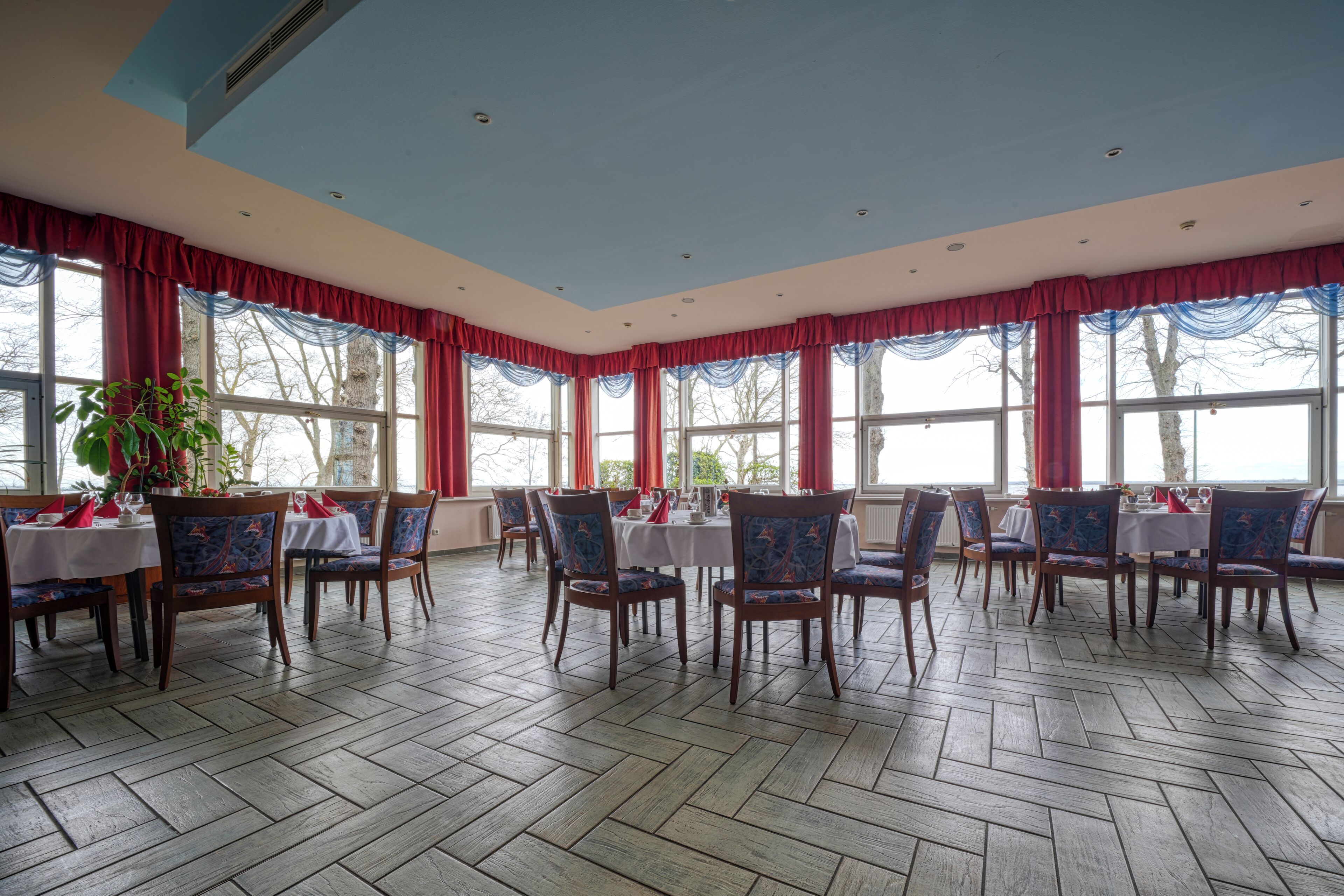A dining area with wooden flooring, red curtains, and multiple tables set for dining.
