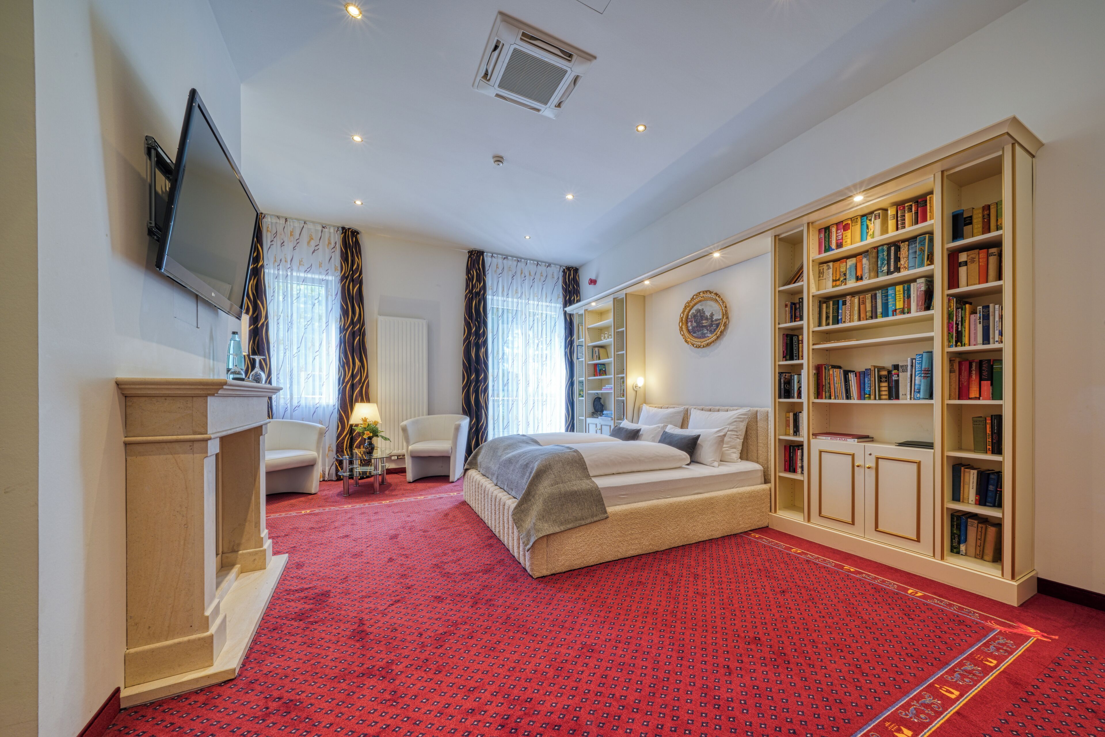 A hotel room with a large bed, a bookshelf, a TV mounted on the wall, and a red carpet.