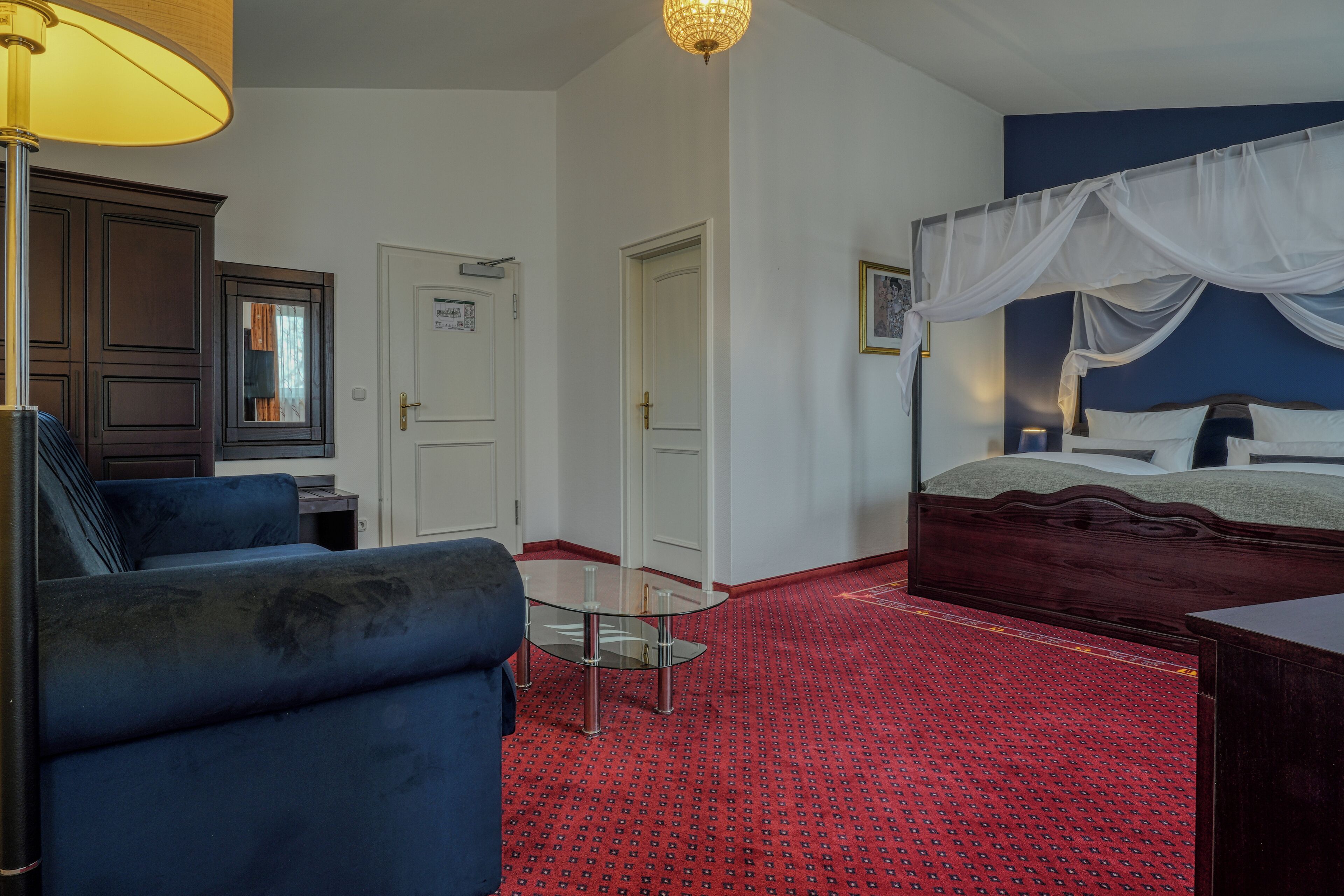 A bedroom with a bed, a glass table, a blue armchair, a wooden cabinet, and a red patterned carpet.