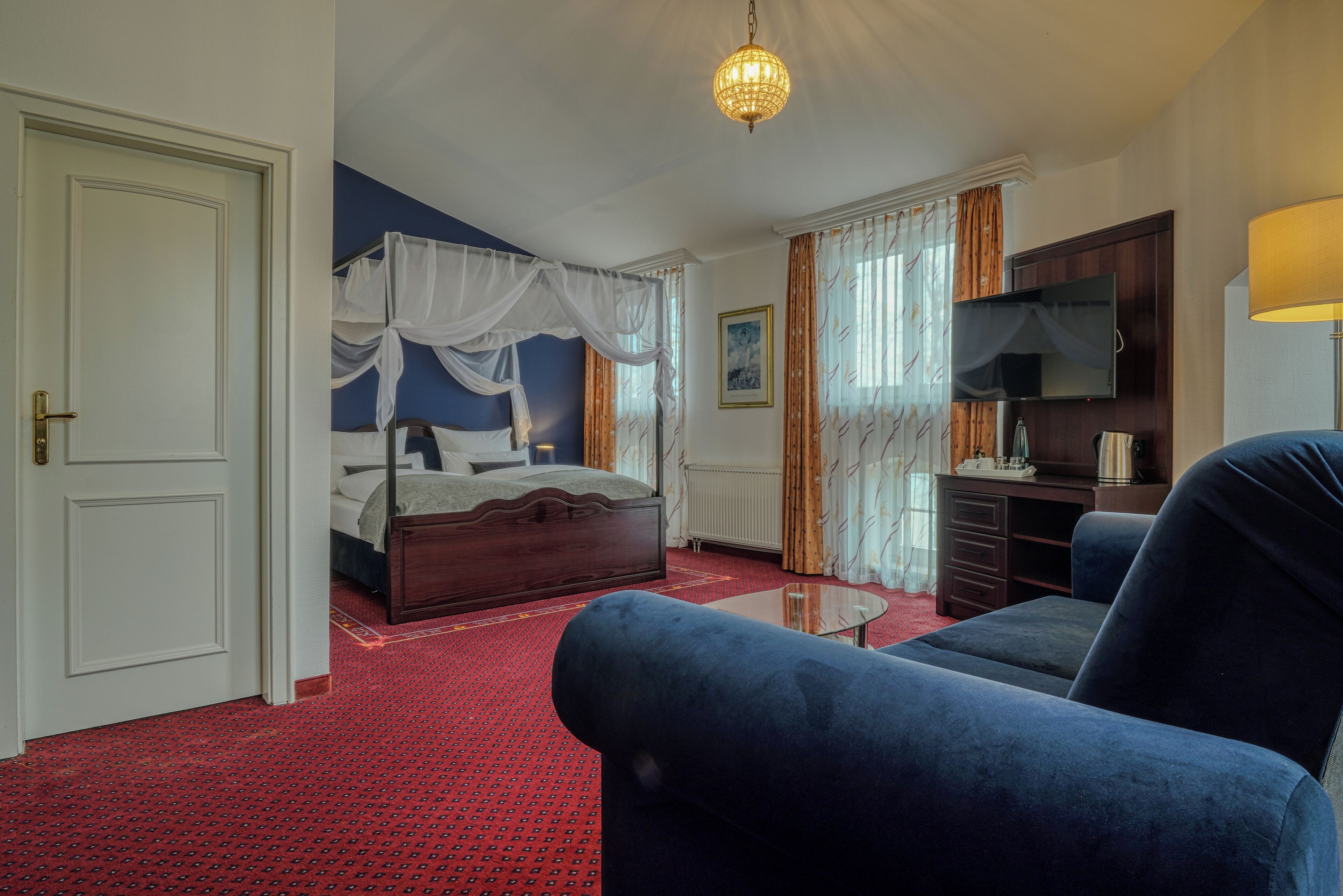 A bedroom with a four-poster bed, a sitting area with a sofa and a coffee table, a dresser, and a television.