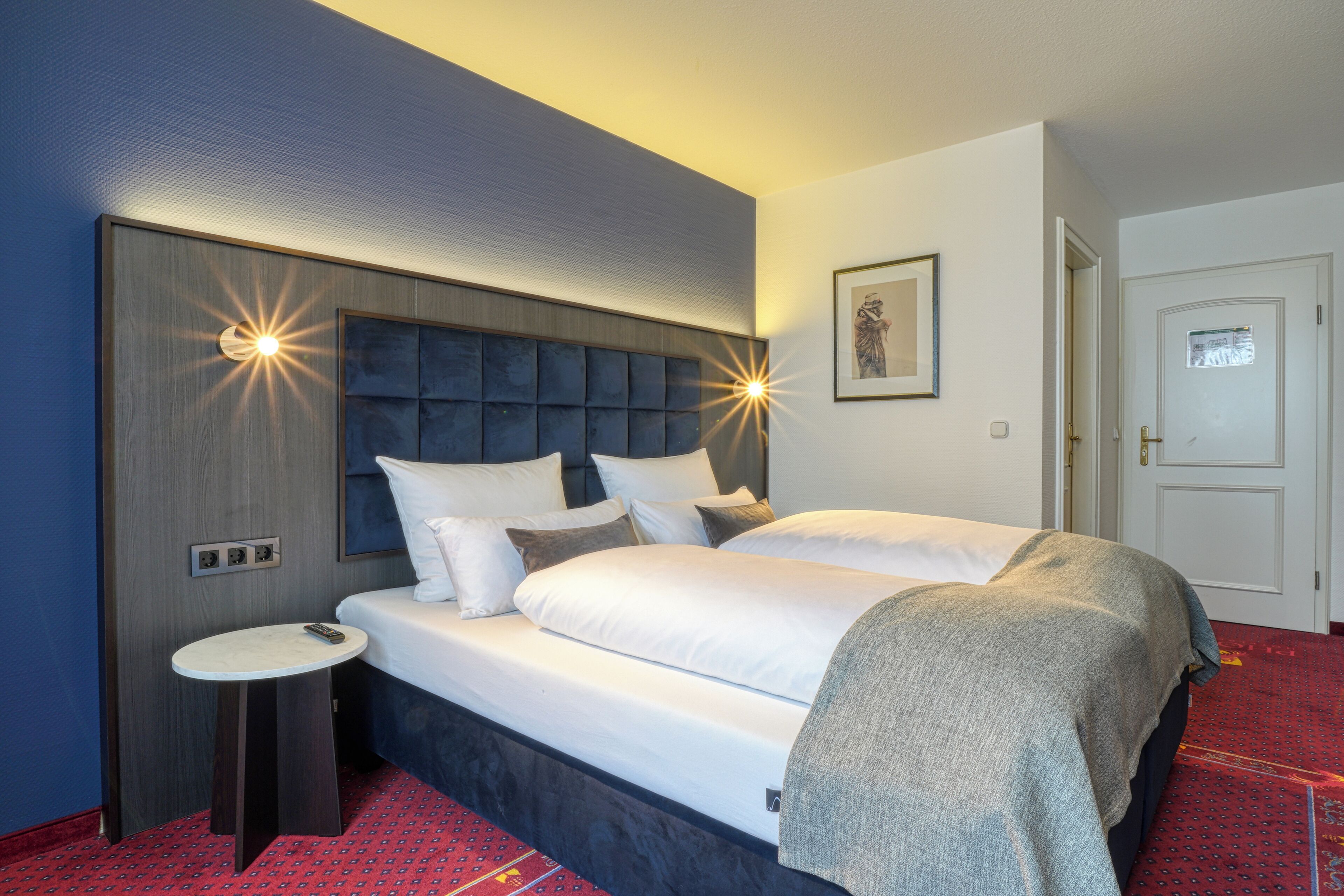 A hotel room with a large bed, a bedside table, a wall-mounted light, and a door.