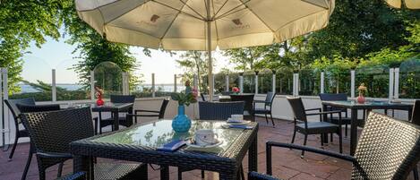 Un patio al aire libre con mobiliario de mimbre, una mesa con cubierta de vidrio y un gran paraguas que ofrece sombra.
