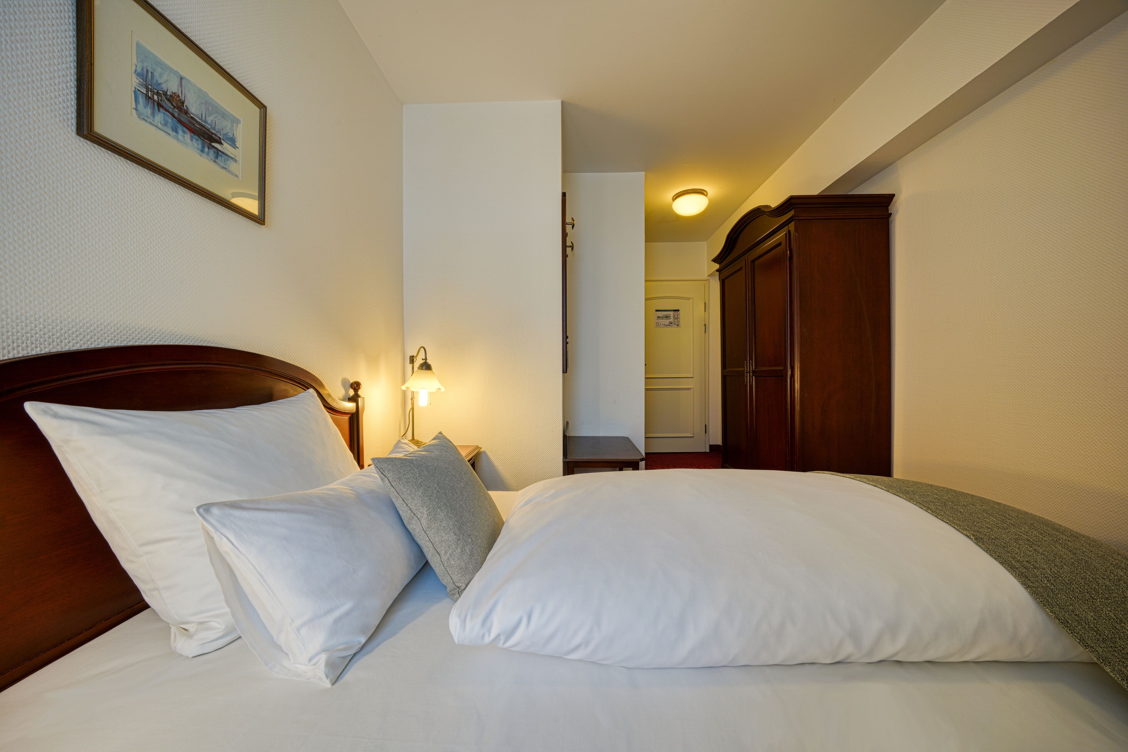 Una cama bien hecha con sábanas y almohadas blancas, un cabecero de madera, una lámpara de noche y un cuadro en la pared.