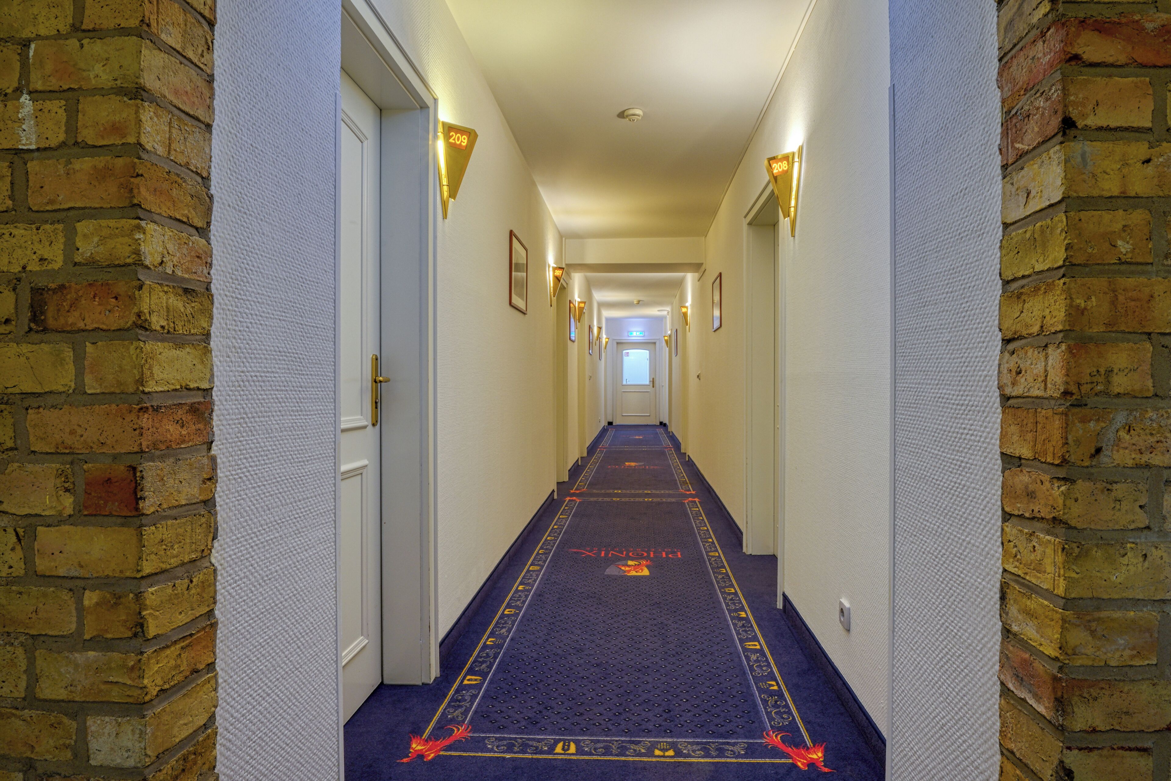 Corredor de hotel com carpete azul, paredes de tijolo e uma porta no final.