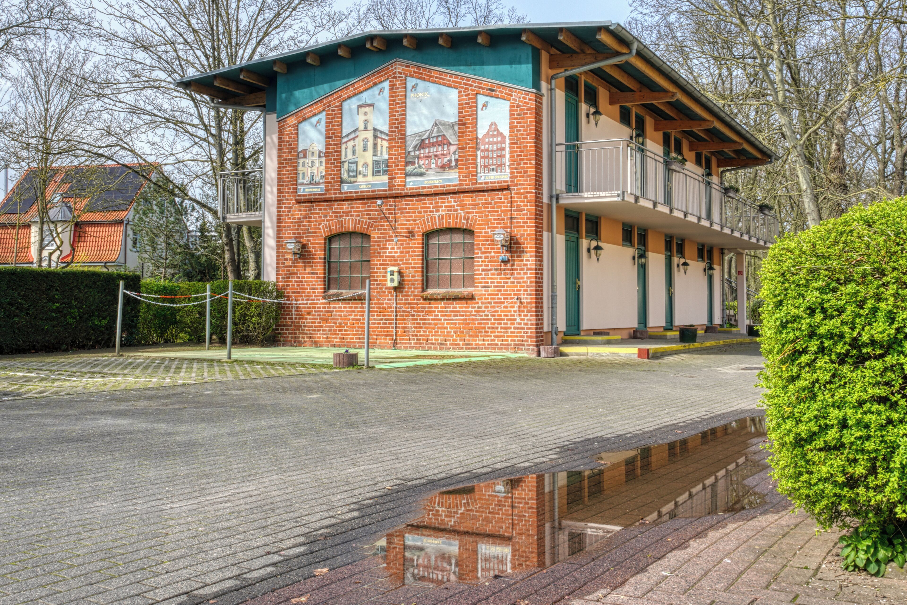 Un edificio in mattoni con un balcone, un riflesso nell'acqua e un siepe sullo sfondo.