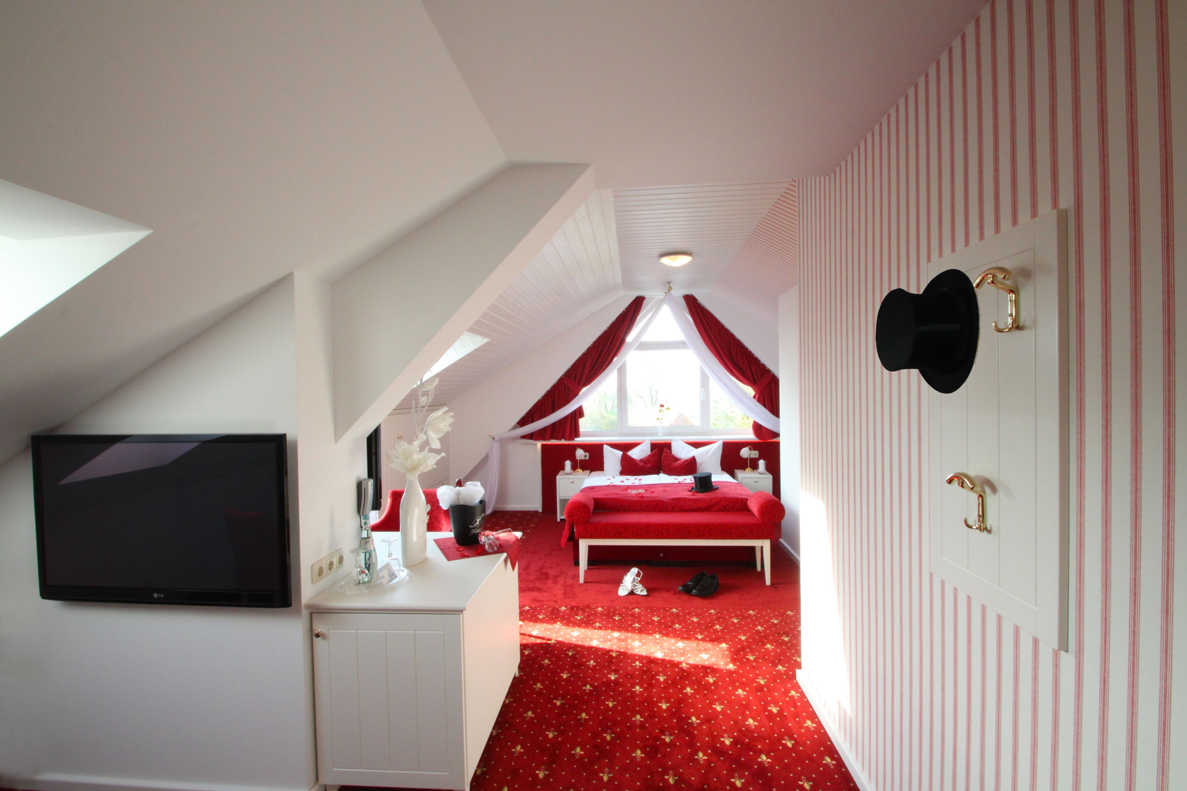 Ein Schlafzimmer mit rotem Teppich, einem Bett mit roter Bettwäsche, einer weißen Kommode, einem Flachbildfernseher und einem Fenster mit roten Vorhängen.